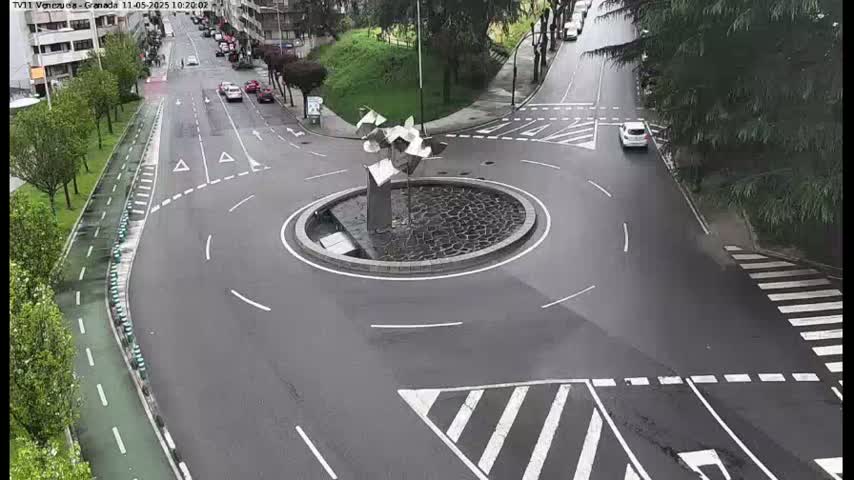 A roundabout with a modern metal sculpture in the center is surrounded by roads with a few cars and a pedestrian crossing, on an overcast day.