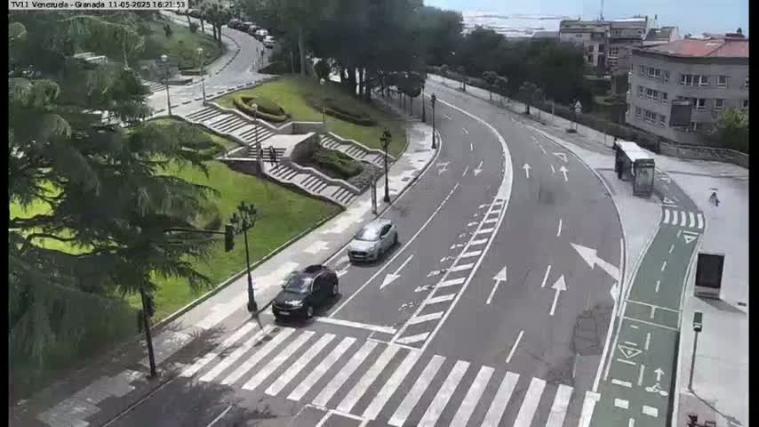 A curved road with two cars and a bus, alongside a pedestrian crossing and a stairway leading to a grassy area, under a partly sunny sky.