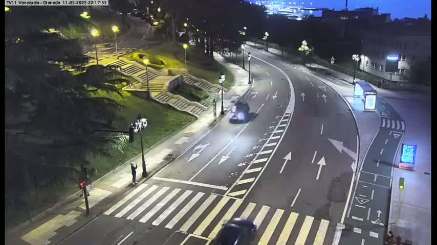 A nighttime, high-angle view shows a curved road with two vehicles and a pedestrian crossing, next to stairs leading up a grassy hill.