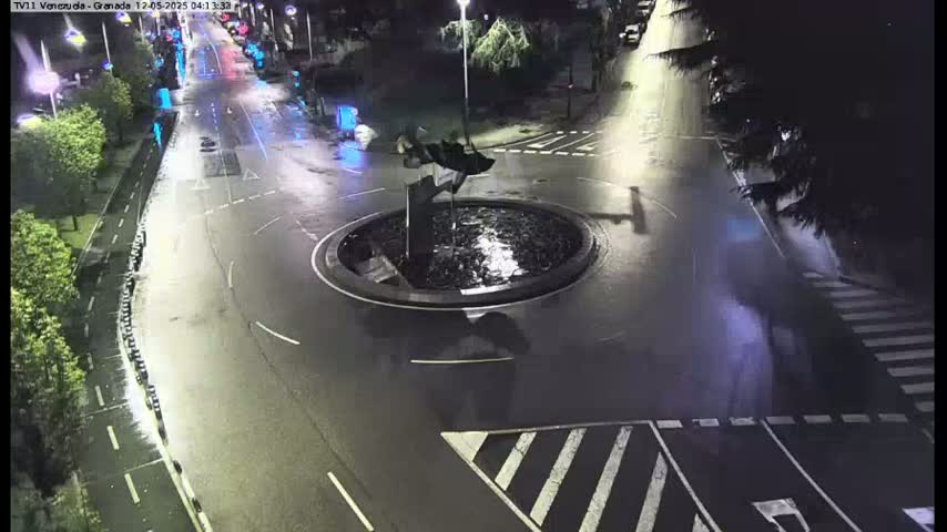A wet roundabout with a dark sculpture in the center is at the intersection of several roadways at night.
