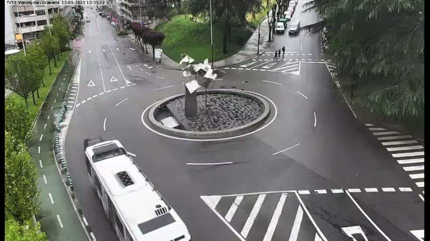A wet roundabout with a modern sculpture in the center, surrounded by roadways with cars and a bus driving on it.
