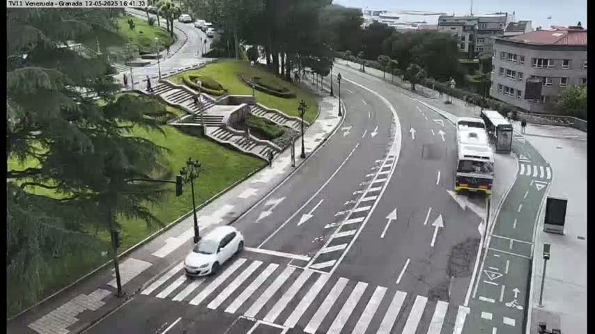 A curved road with a crosswalk and a bike lane passes by a grassy area with stairs, a few pedestrians, and two buses and a car are visible on the road.