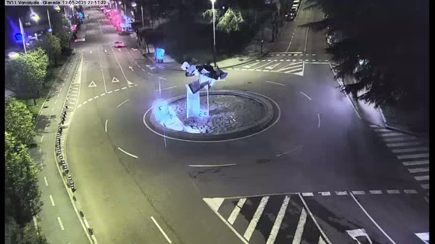 A nighttime aerial view shows a roundabout with a modern sculpture in its center, surrounded by roadways with minimal traffic.