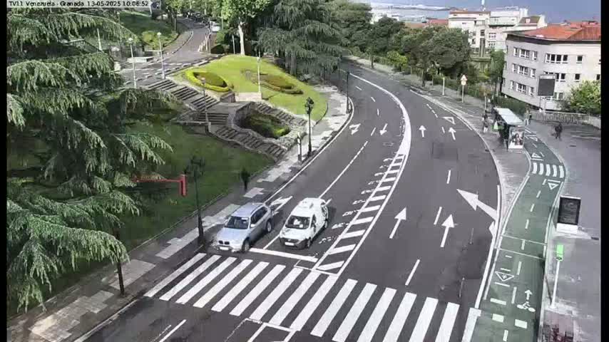 A curved roadway with crosswalks and a bike lane, alongside a park-like area with stairs and lush greenery, under a mostly cloudy sky.
