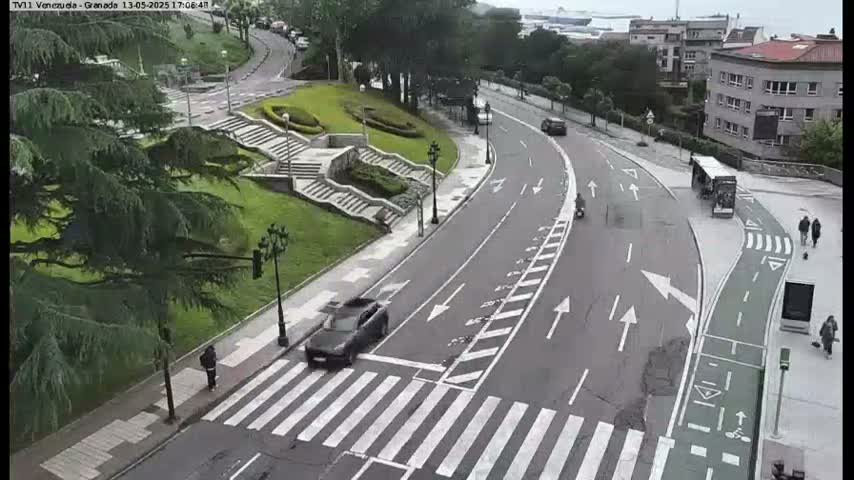 A curved road with pedestrian crossings and bike lanes winds through a city park on an overcast day, with a few cars, pedestrians, and a bus visible.