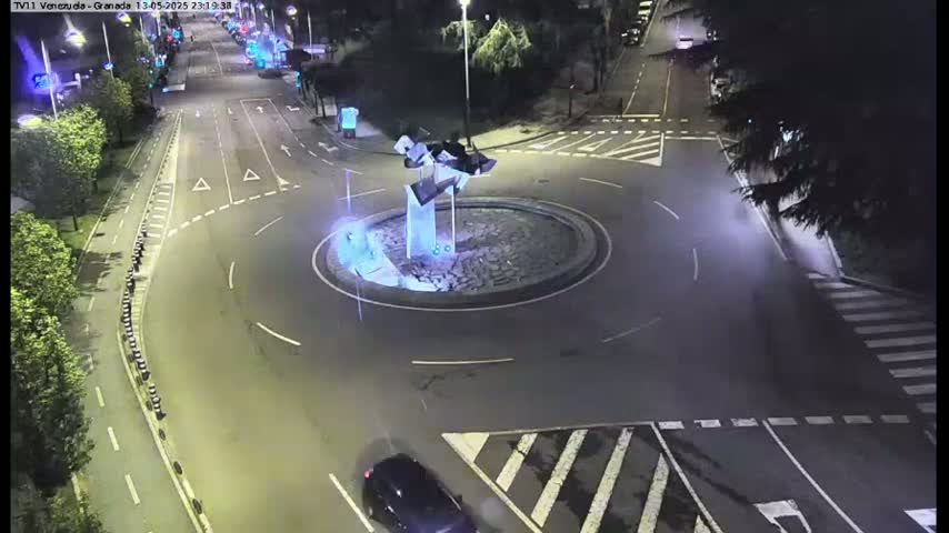 A nighttime aerial view shows a roundabout with a modern sculpture in the center, surrounded by roads and cars.