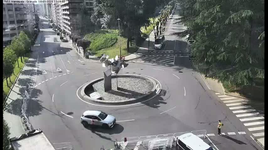 A sunny day shows a roundabout with a modern sculpture in the center, surrounded by a city street with several cars and a pedestrian.