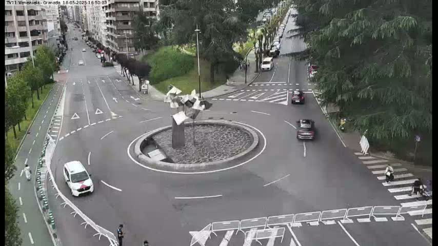An overcast day shows a roundabout with several cars, pedestrians crossing a crosswalk, and a sculpture in the center, all situated within a city setting.