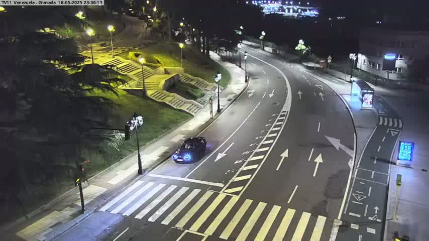 A dark-colored car is stopped at a crosswalk on a curving, well-lit road at night.