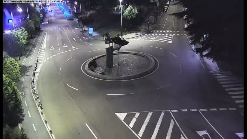 A nighttime aerial view shows a traffic circle with a dark sculpture in the center, surrounded by roads with minimal traffic.