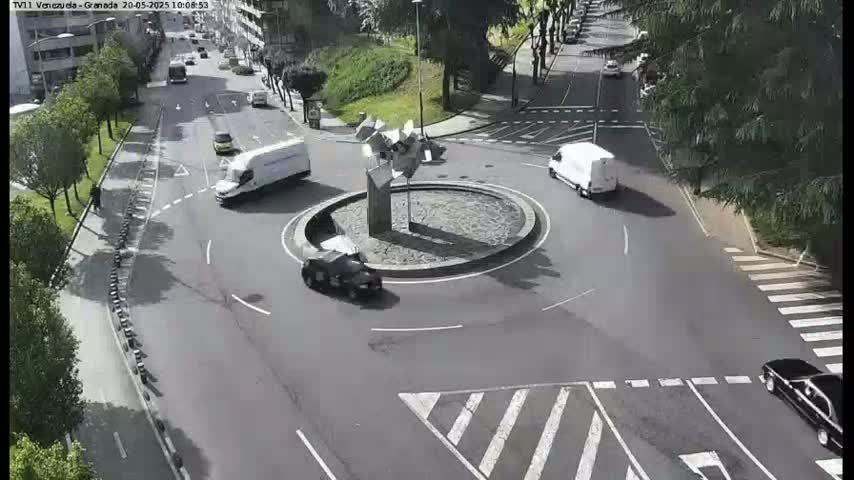 Several vehicles navigate a roundabout in a sunny city setting, with one car passing a sculpture at its center.