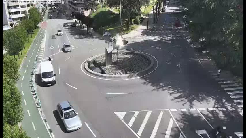 A sunny day shows several cars navigating a roundabout with a central sculpture, surrounded by trees and a pedestrian crossing.