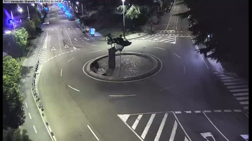 A nighttime aerial view of a roundabout with a dark sculpture in the center, surrounded by roads and pedestrian crossings.