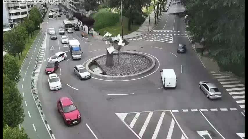 Several cars navigate a roundabout on a sunny day, with a modern sculpture at its center.