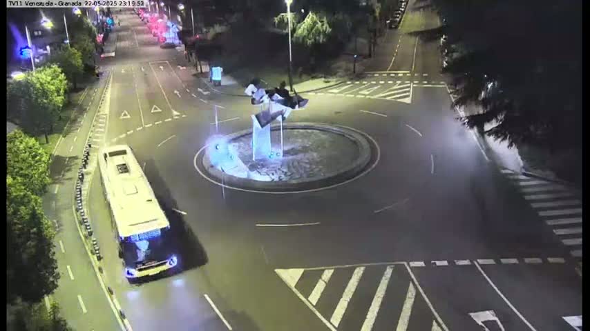 A large, light-colored bus approaches a roundabout at night, illuminated by streetlights, with a modern sculpture in the center.