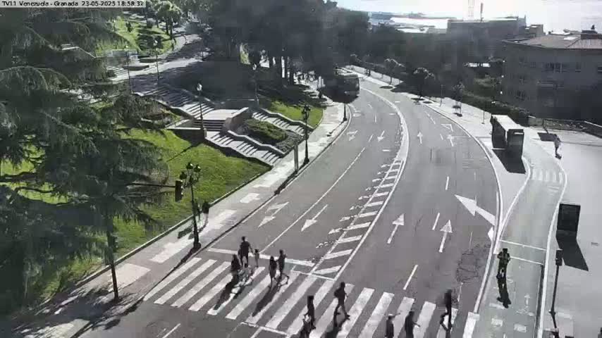 A sunny day shows a roadway curving through a city with pedestrians crossing a crosswalk and a bus driving down the street, next to green spaces with stairs and trees.