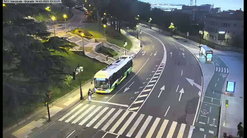A long, curved road at night is seen with a bus stopped at a crosswalk, another bus further down the road, and pedestrians crossing.