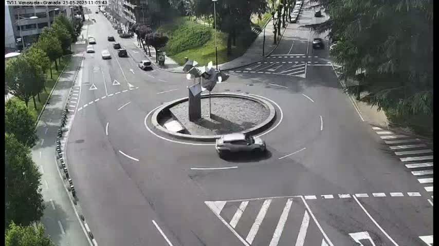 A silver car drives through a roundabout in the center of a roadway on a partly sunny day.