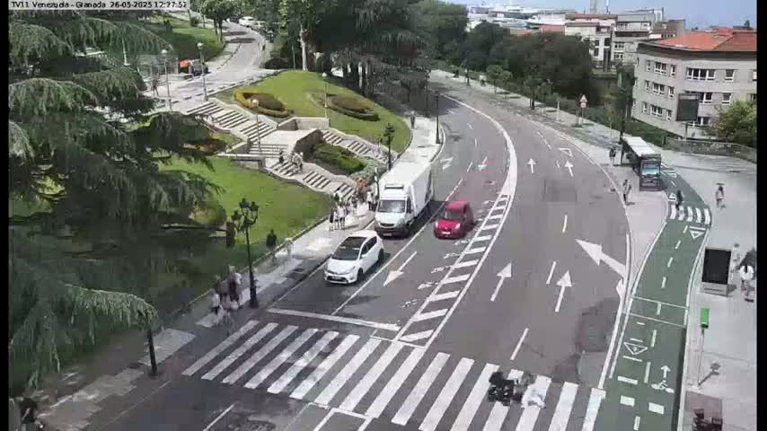 A sunny day shows a curved road with cars and pedestrians, a crosswalk, and a bus stop near a grassy area with stairs and trees.