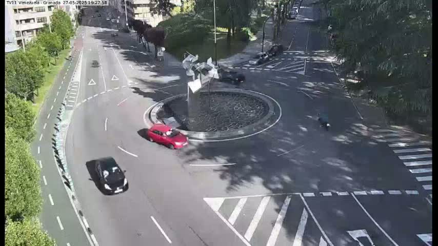 A sunny day shows a roundabout with several cars and a motorcyclist navigating it, surrounded by trees and a roadway with a bike lane.