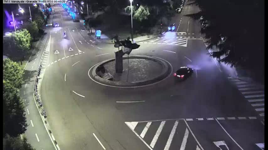 A nighttime, overhead view of a roundabout with a dark-colored car driving through it and surrounding streets with minimal traffic.