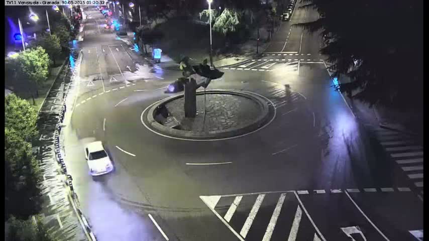 A white car is performing a turn in a roundabout at night, leaving tire marks on the wet asphalt.