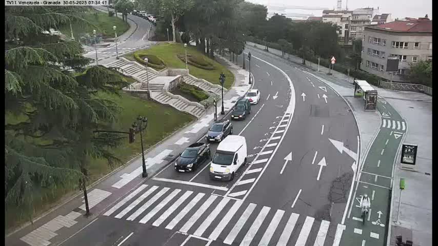 A curved road with several cars and a van at a crosswalk, near a stairway leading to a grassy area with trees and buildings in the background, on an overcast day.