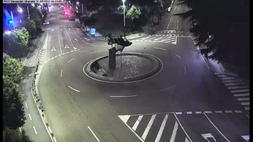 A nighttime, aerial view of a roundabout with a dark sculpture in the center, surrounded by roads with white markings.