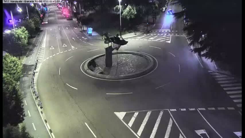 A nighttime aerial view of a traffic circle with a sculpture in the center, surrounded by roads and trees.