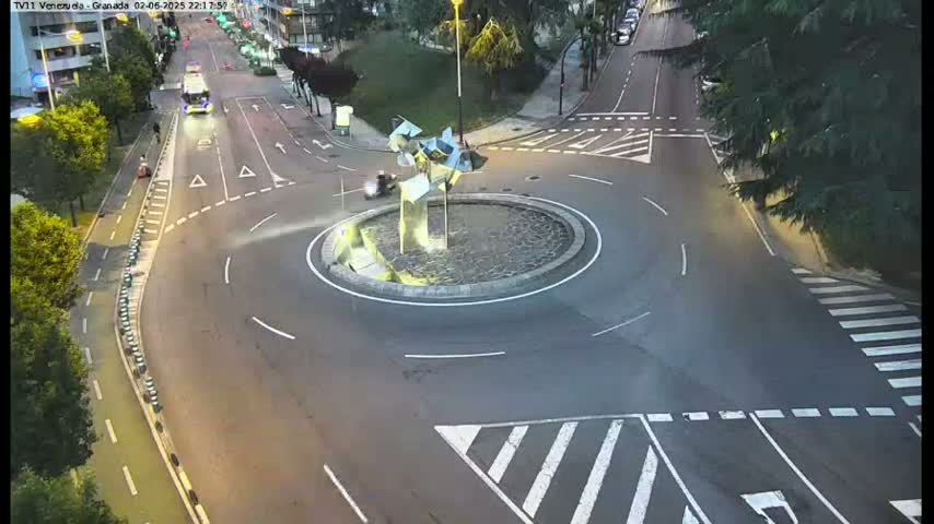 A high-angle, nighttime view of a roundabout with a modern sculpture in the center, surrounded by roads and pedestrian crossings.