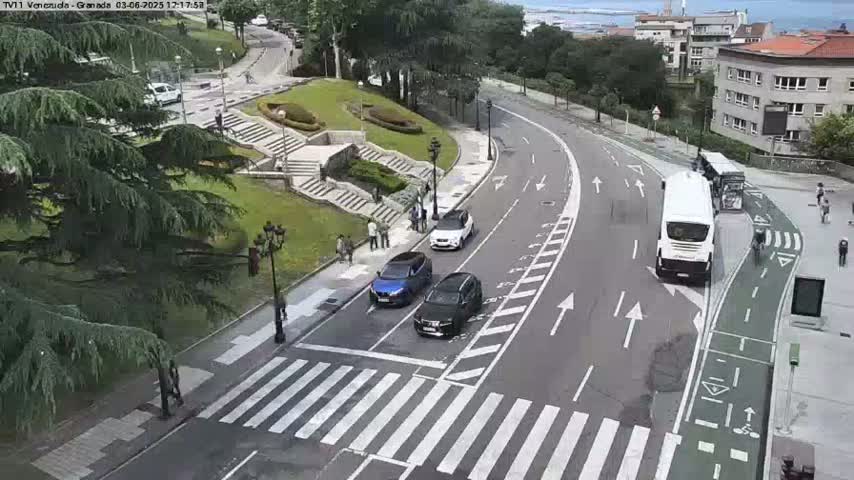 A curving road with several cars and a bus, a pedestrian crossing, and a stairway leading to a grassy area with trees and buildings in the background on a sunny day.