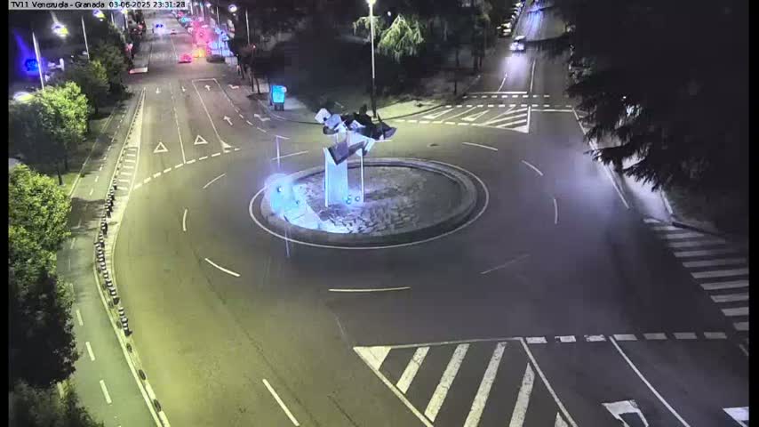 A nighttime aerial view shows a roundabout with a modern sculpture in the center, surrounded by roads with minimal traffic.
