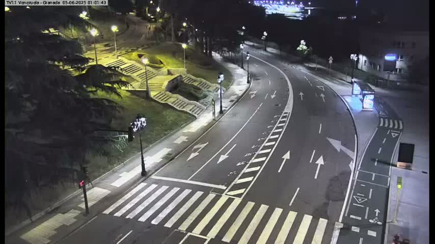 A nighttime, high-angle view of a mostly empty roadway curving through a park-like setting with stairs and streetlights, under clear skies.