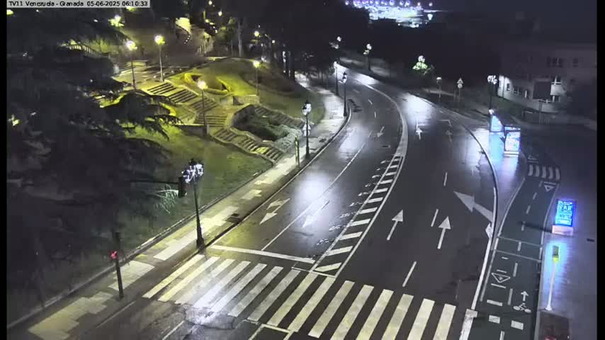 A wet, nighttime street scene shows a curving road with pedestrian crossings, streetlights, and a staircase leading up to a grassy area with trees.