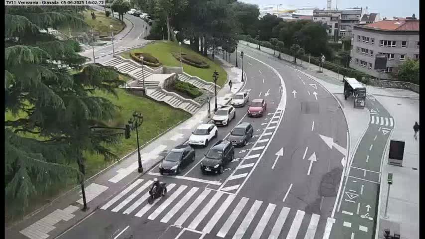 A curved road with several cars and a motorcycle at a crosswalk, alongside a set of stairs leading down to a grassy area, all under an overcast sky.