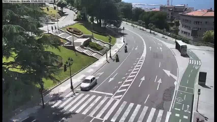 A sunny day reveals a winding road curving through a landscaped area with stairs, greenery, and a few pedestrians and vehicles.