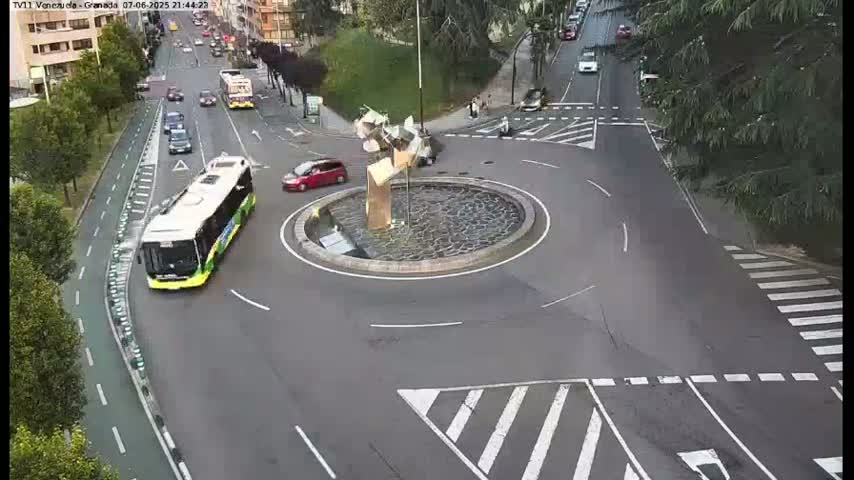 A city roundabout with a sculpture in the center, surrounded by cars and a bus, on a partly sunny day.