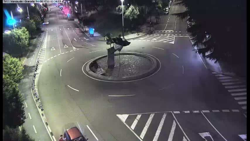 A nighttime aerial view shows a roundabout with a dark sculpture in the center, surrounded by roadways and pedestrian crosswalks.