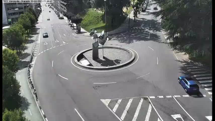A sunny day shows a roundabout in the center of a road intersection, with a few cars visible and lush green trees lining the streets.