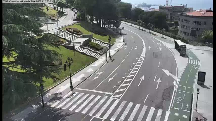 A winding road with a pedestrian crossing curves through a park-like area with stairs and lush green landscaping on a sunny day.