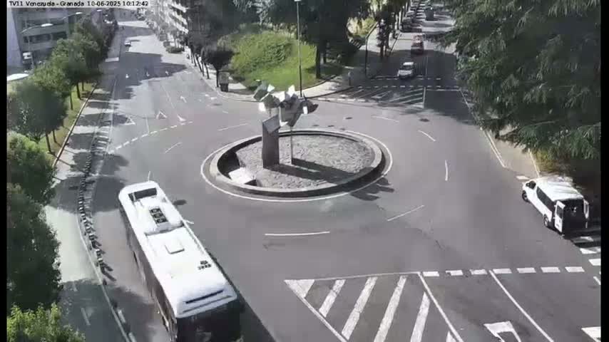 A white bus approaches a roundabout with a modern sculpture in the center, under a sunny sky.