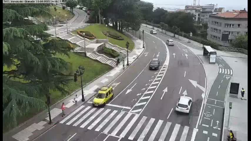 A curved roadway with several cars and pedestrians, situated beside a grassy area with stairs and trees, on an overcast day.