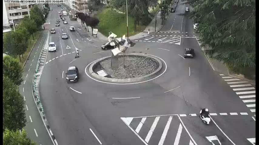 A roundabout with a modern sculpture in the center, surrounded by several cars and a scooter, on a mostly sunny day.