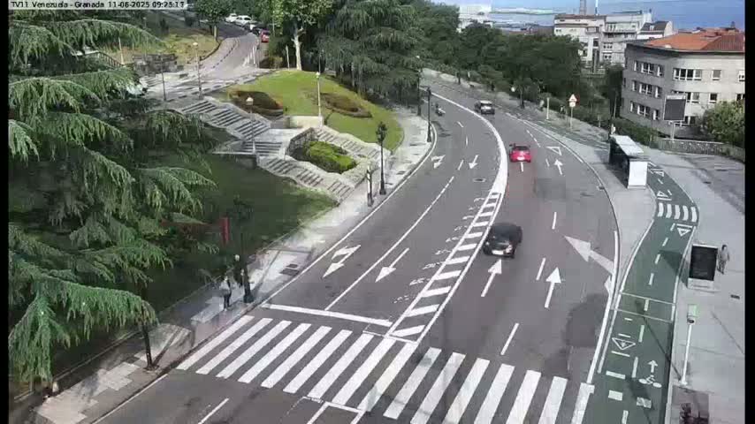 A paved road curves through a landscaped area with a pedestrian crossing, several cars are driving on the road, and a few people are walking near a crosswalk on a partly sunny day.