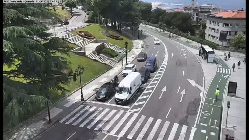 A sunny day overlooking a curving road with several cars and a pedestrian crossing, near a park with stone steps and lush greenery.