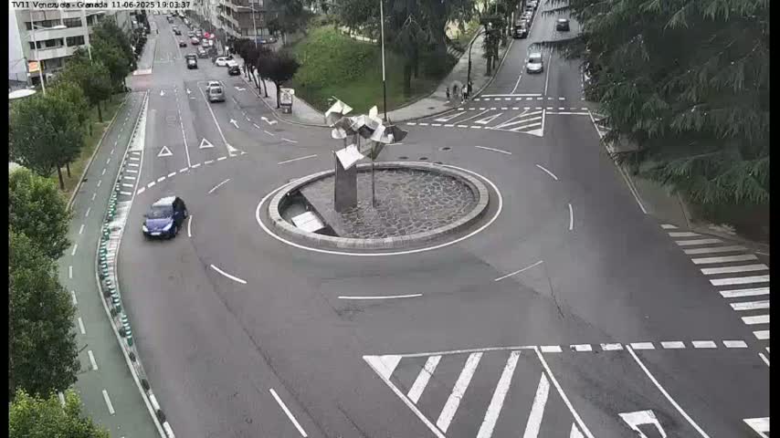 A roundabout with a modern metal sculpture in the center, surrounded by roads with several cars and a bicycle lane, on an overcast day.