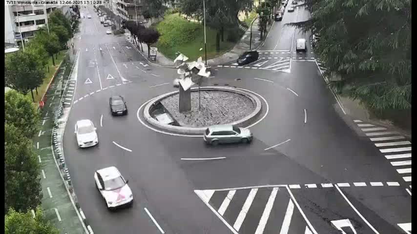 Several cars navigate a wet roundabout centered around a modern sculpture.