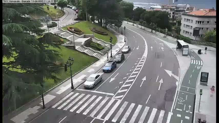 A curving road with several parked cars passes by a grassy area with stairs leading to a building, all under overcast skies.