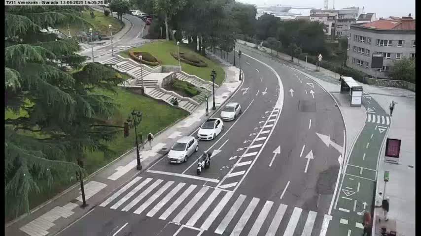 A curved road with several cars and a pedestrian crossing, near a staircase leading to a grassy area and some buildings, under overcast skies.