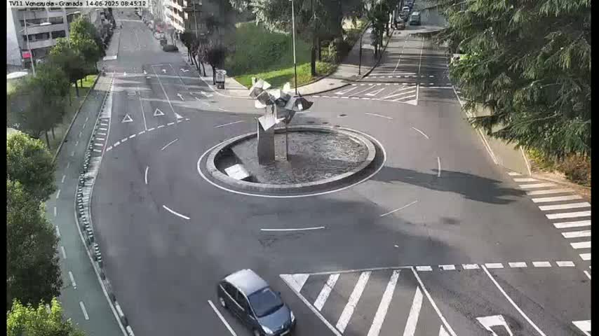 A roundabout with a modern metallic sculpture in the center is situated on a paved road surrounded by trees and buildings under a sunny sky.